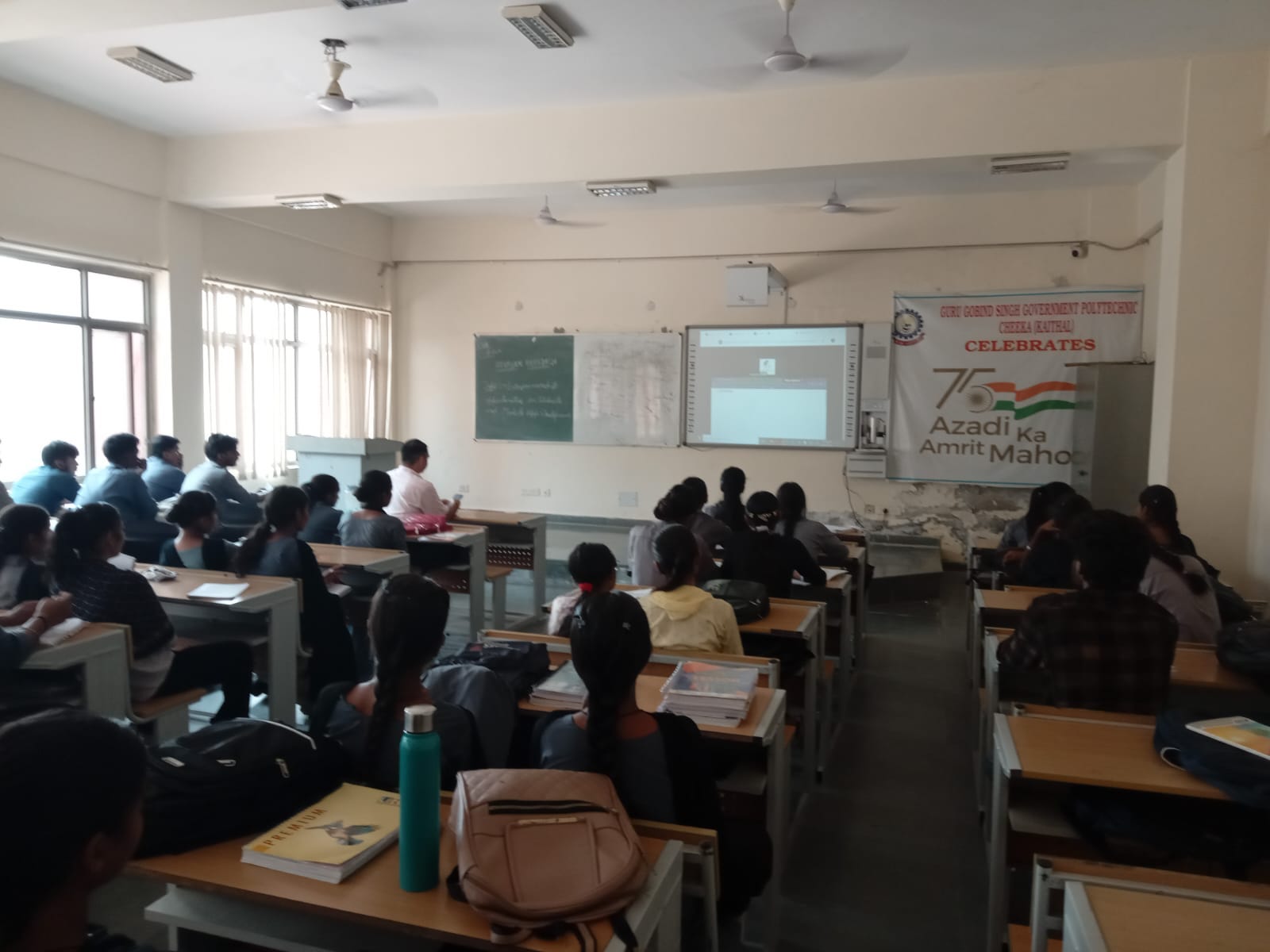 Students in a smart classroom facing an interactive display board, with a college banner on the wall.