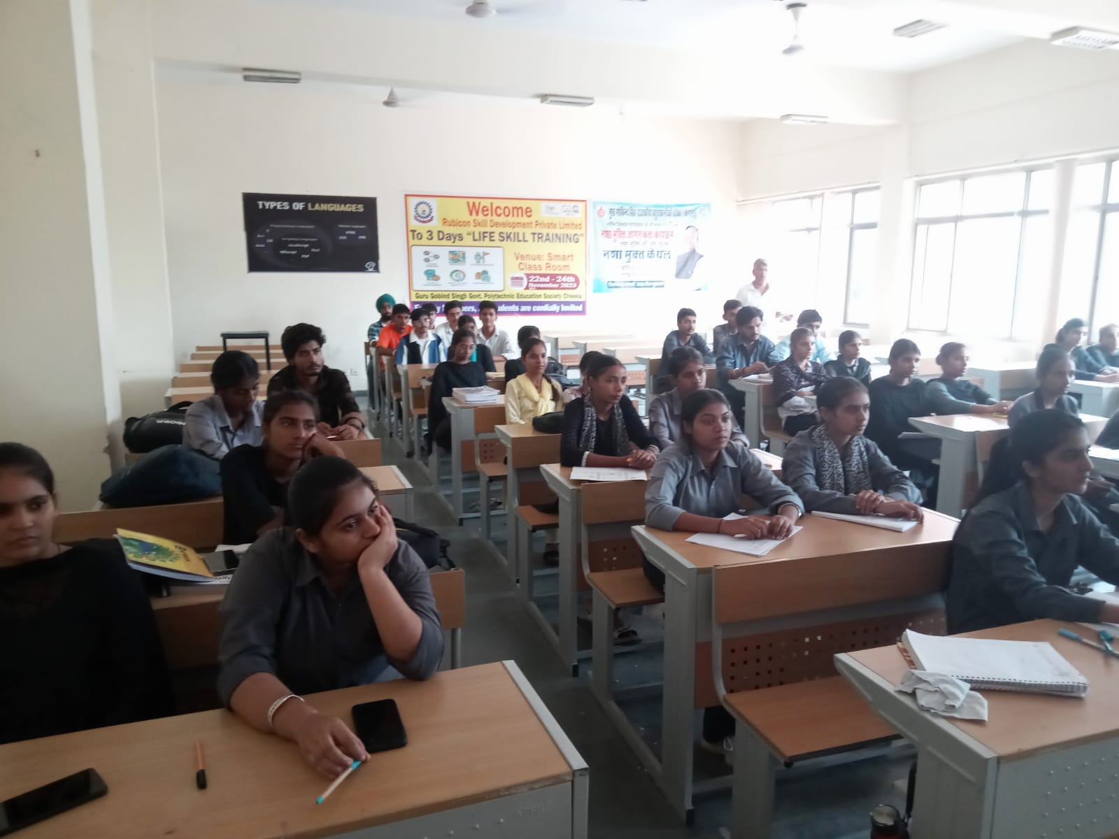 Students facing the front of a classroom, with a programming-languages poster and training banners on the wall.